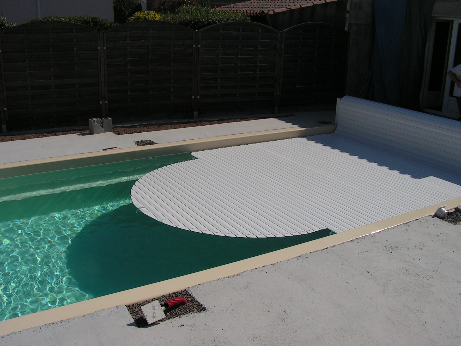Pool with a white corrugated cover partially rolled back on a concrete deck; dark wooden privacy fence in the background.