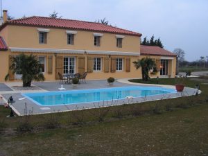 Two-story yellow villa with a red-tiled roof, wooden shutters, and striped awnings beside a rectangular blue pool.",